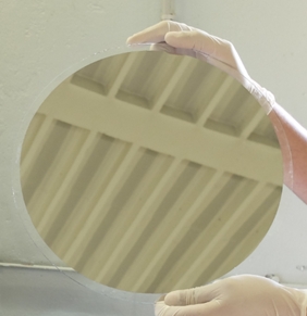 Photo of gloved hands holding a Ø300mm glass plate with a mirror like deposit reflecting the ceiling, realized by multi beam sputtering